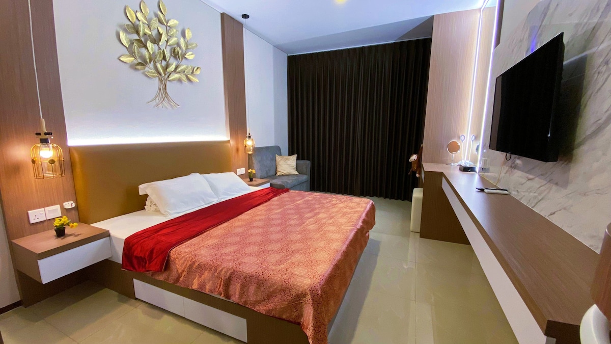 A modern studio room features a spacious bed dressed in a red and gold patterned coverlet. A cozy seating area is provided by a gray armchair. Decorative wall art enhances the ambiance, while large curtains partially block natural light, contributing to a comfortable atmosphere.