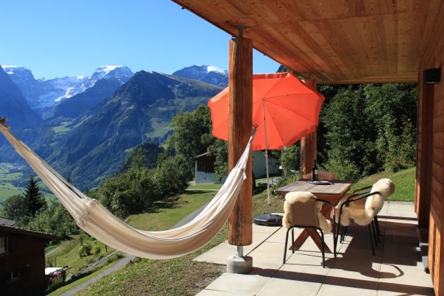 A covered outdoor area features a hammock and a dining set beneath a vibrant orange umbrella. The mountains of Glarner are visible in the background, creating a serene and scenic environment.