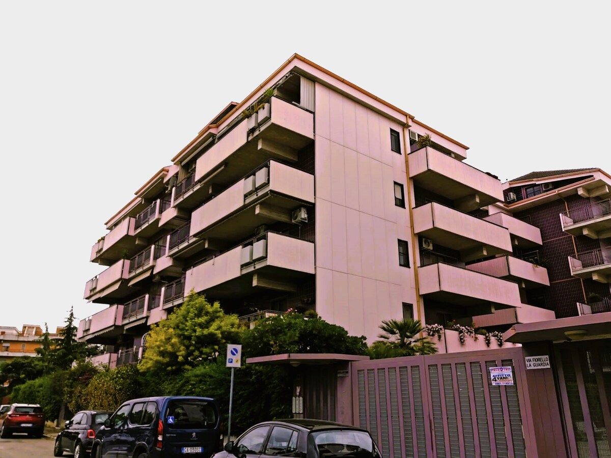 A modern four-story building features multiple balconies with greenery, enhancing the exterior appeal. The structure is flanked by trees and shrubs, contributing to a serene environment. Several vehicles are parked along the street, providing a glimpse of the community's accessibility.