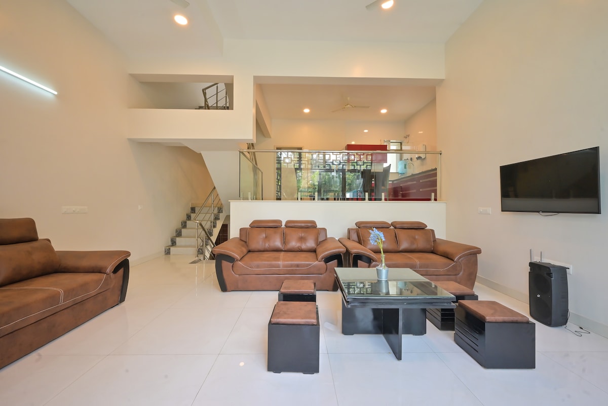 A spacious living area features modern leather sofas arranged around a glass coffee table. Large windows allow natural light to fill the room, while a sleek television is mounted on the wall. A staircase is visible in the background, adding to the open layout.