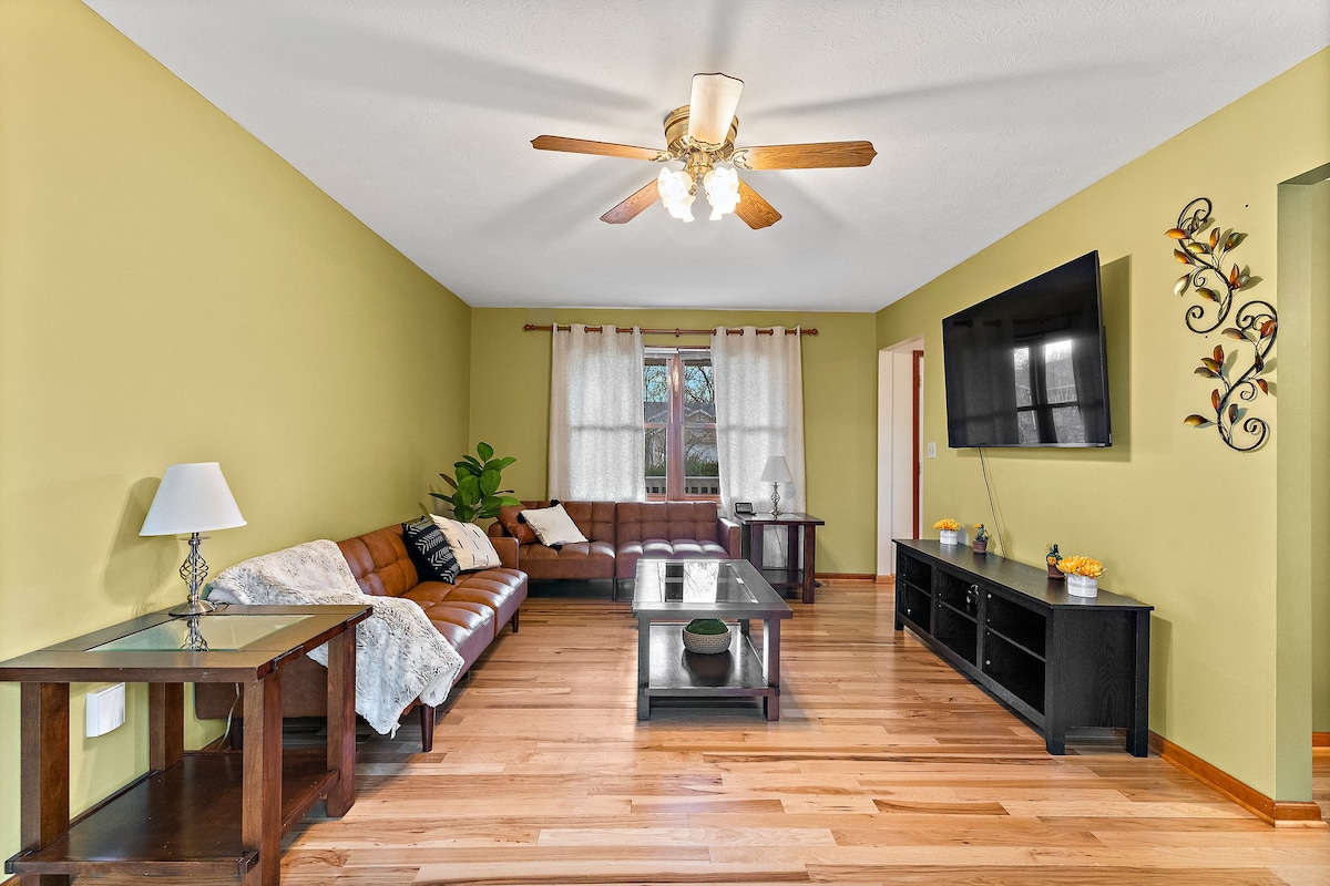 A spacious living room features a combination of wooden flooring and a light green wall. A comfortable brown sofa is positioned against the wall, accompanied by a glass-topped coffee table. Natural light enters through the window, and a flat-screen TV is mounted nearby.