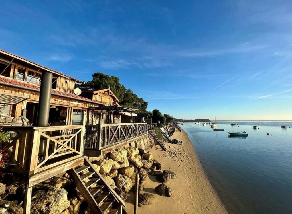 A coastal cabin extends towards the sandy beach, featuring a wooden deck with railings. Steps lead down to the water's edge where small boats are anchored. The scene is framed by trees and a clear blue sky, contributing to a serene seaside setting.
