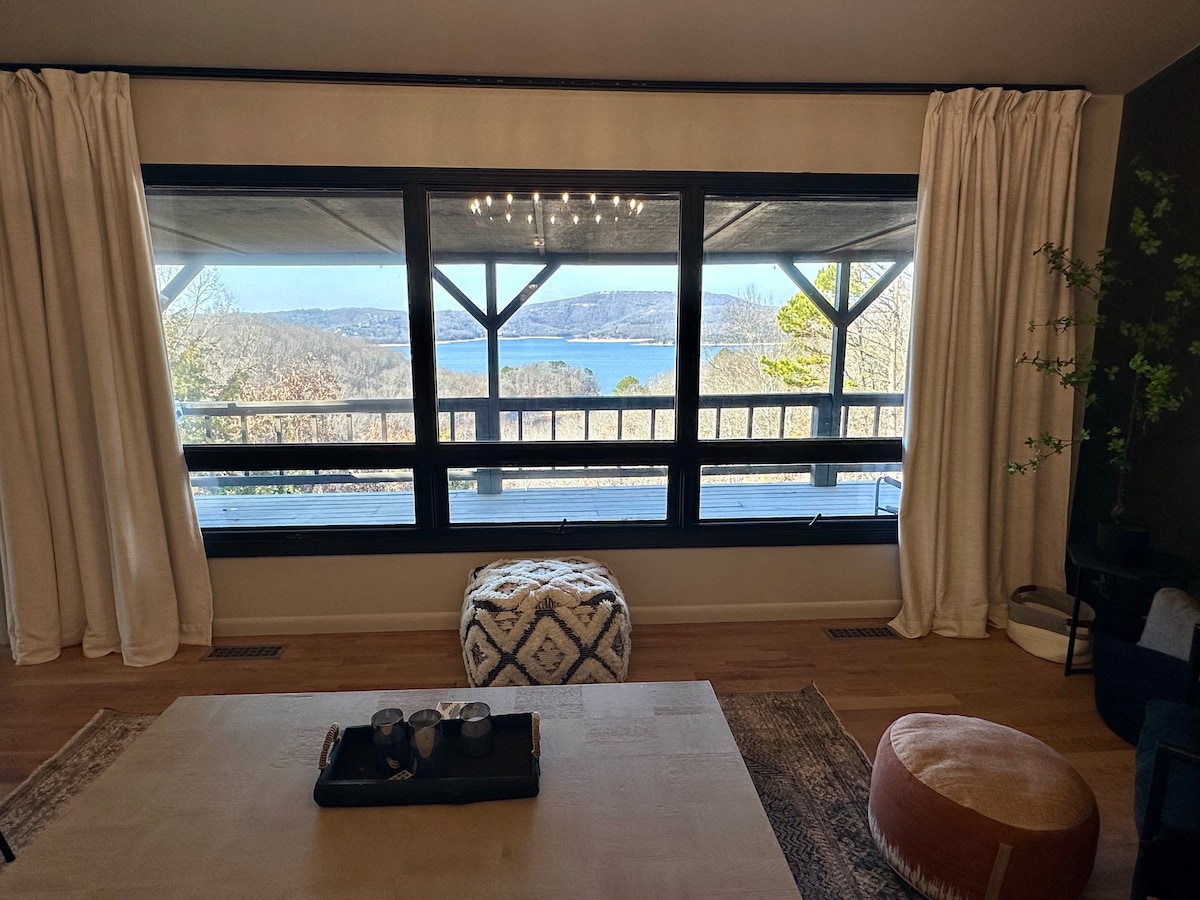 Large windows offer a panoramic view of the surrounding landscape and water, framed by light, flowing drapes. A low coffee table sits in the center of the room, with decorative items arranged atop it. Natural light illuminates the space, enhancing its openness.