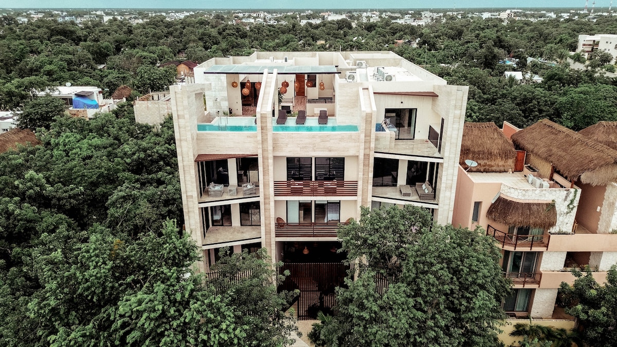 Aerial view of a modern three-story apartment building surrounded by lush greenery. The structure features balconies with lounge chairs and a rooftop terrace, highlighted by a swimming pool. The architectural design integrates natural elements, creating a harmonious blend with the landscape.