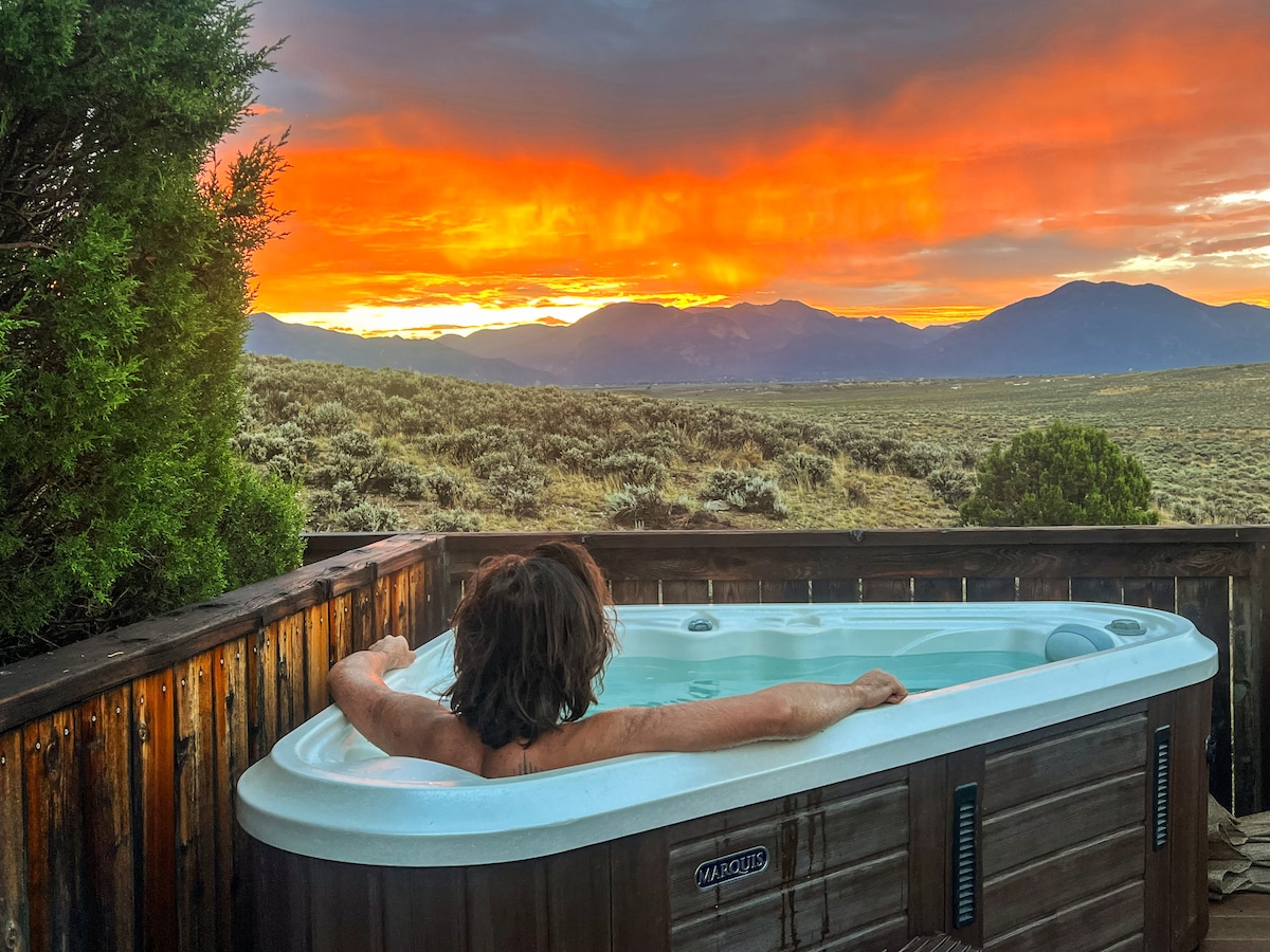 A private hot tub is positioned on a wooden deck, overlooking expansive views of mountains and desert landscapes. A person relaxes in the tub, framed by greenery, as a vibrant sunset casts warm hues across the sky.