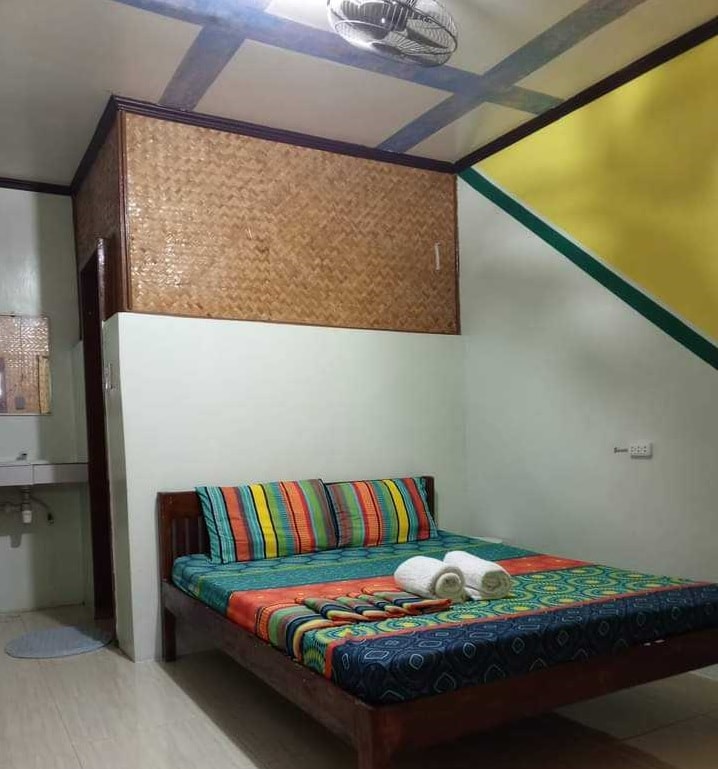 A cozy sleeping area features a wooden bed adorned with a vibrant patterned mattress and matching pillows. Soft, rolled towels are placed on the bed. The walls are painted in light tones with a striking yellow accent, complemented by textured bamboo paneling.