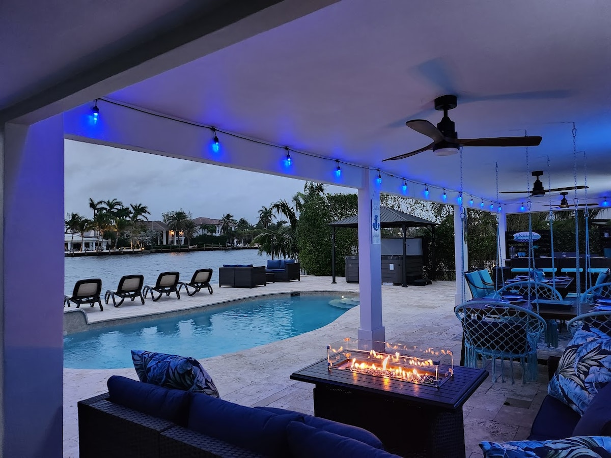 An outdoor area features a heated pool surrounded by lounge chairs, with palm trees visible in the background. A seating arrangement with a fire table is under soft blue lighting. Water views can be seen beyond the pool, creating a tranquil setting.