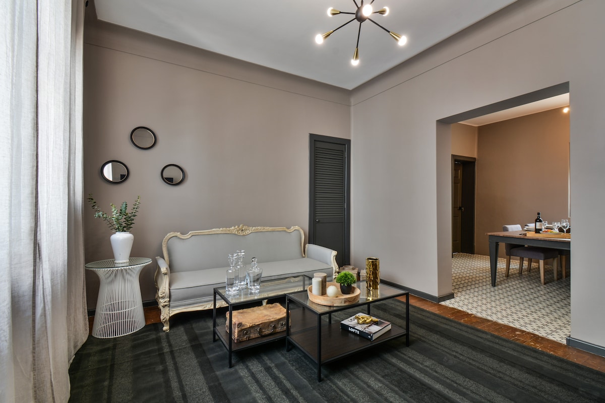 A stylish living area features a classic sofa and a glass coffee table placed on a contemporary rug. Decorative elements like a plant and candles are arranged on the table. Soft natural light filters in through large curtains, highlighting the refined neutral color palette.
