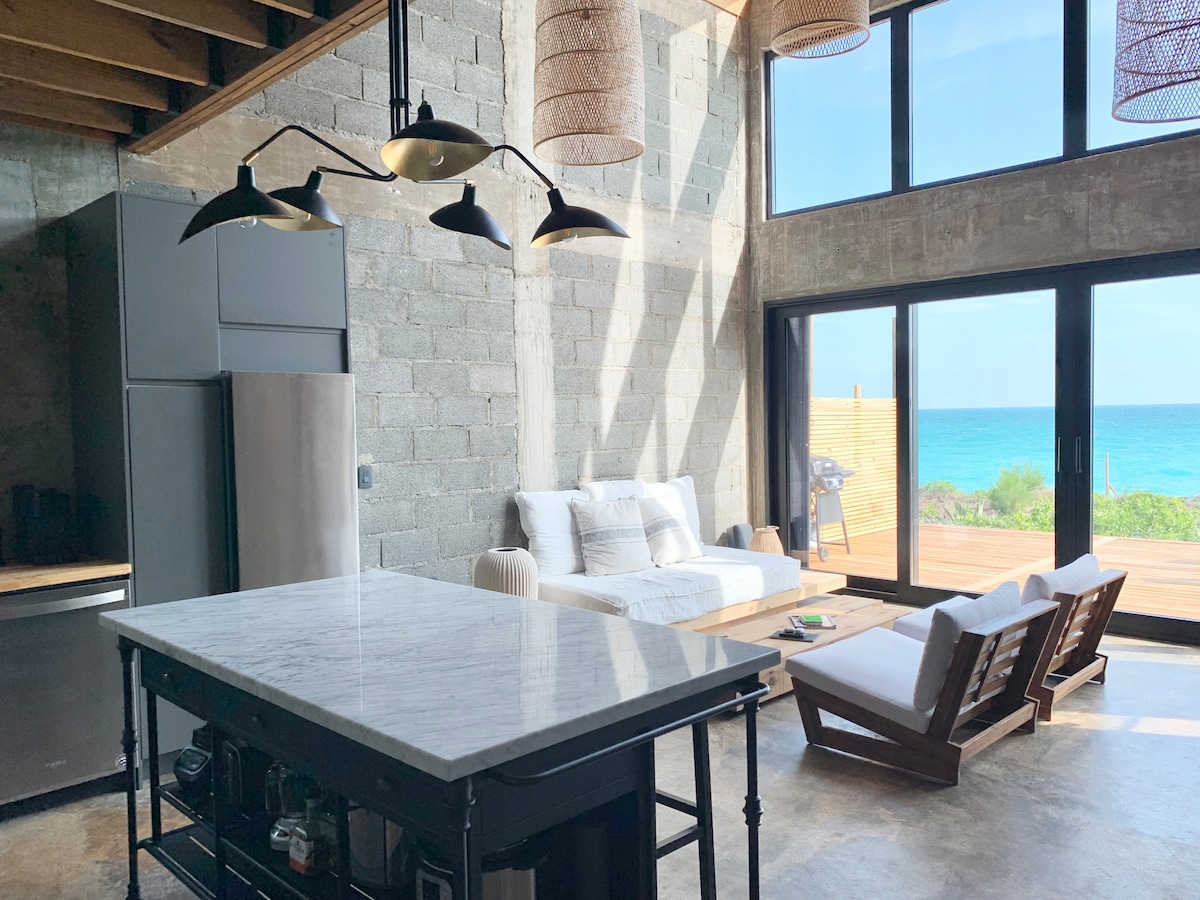 The great room features a spacious marble-top island with seating, adjacent to a cozy living area furnished with a sofa and two lounging chairs. Large industrial-style windows frame a stunning ocean view, allowing natural light to fill the space.