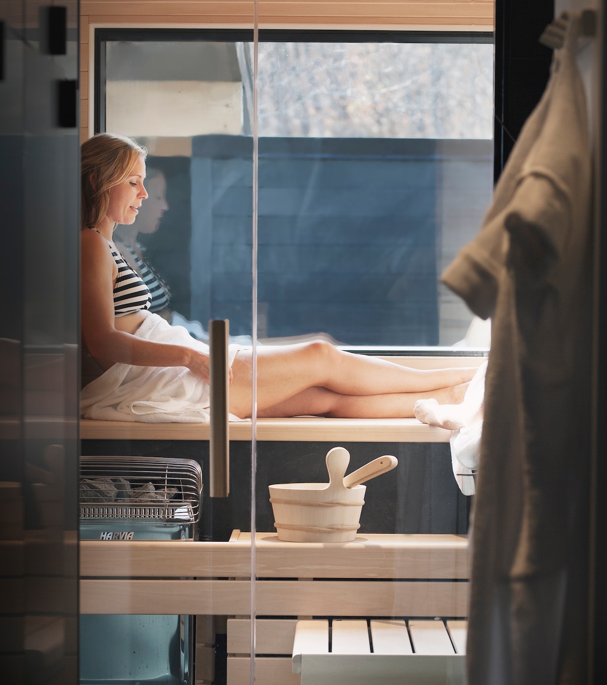A sauna is shown with a relaxed individual seated on a wooden bench, enjoying the warm ambiance. Natural light filters through a window, illuminating the serene space. A bowl and pestle are situated nearby, adding a calming touch to the environment.