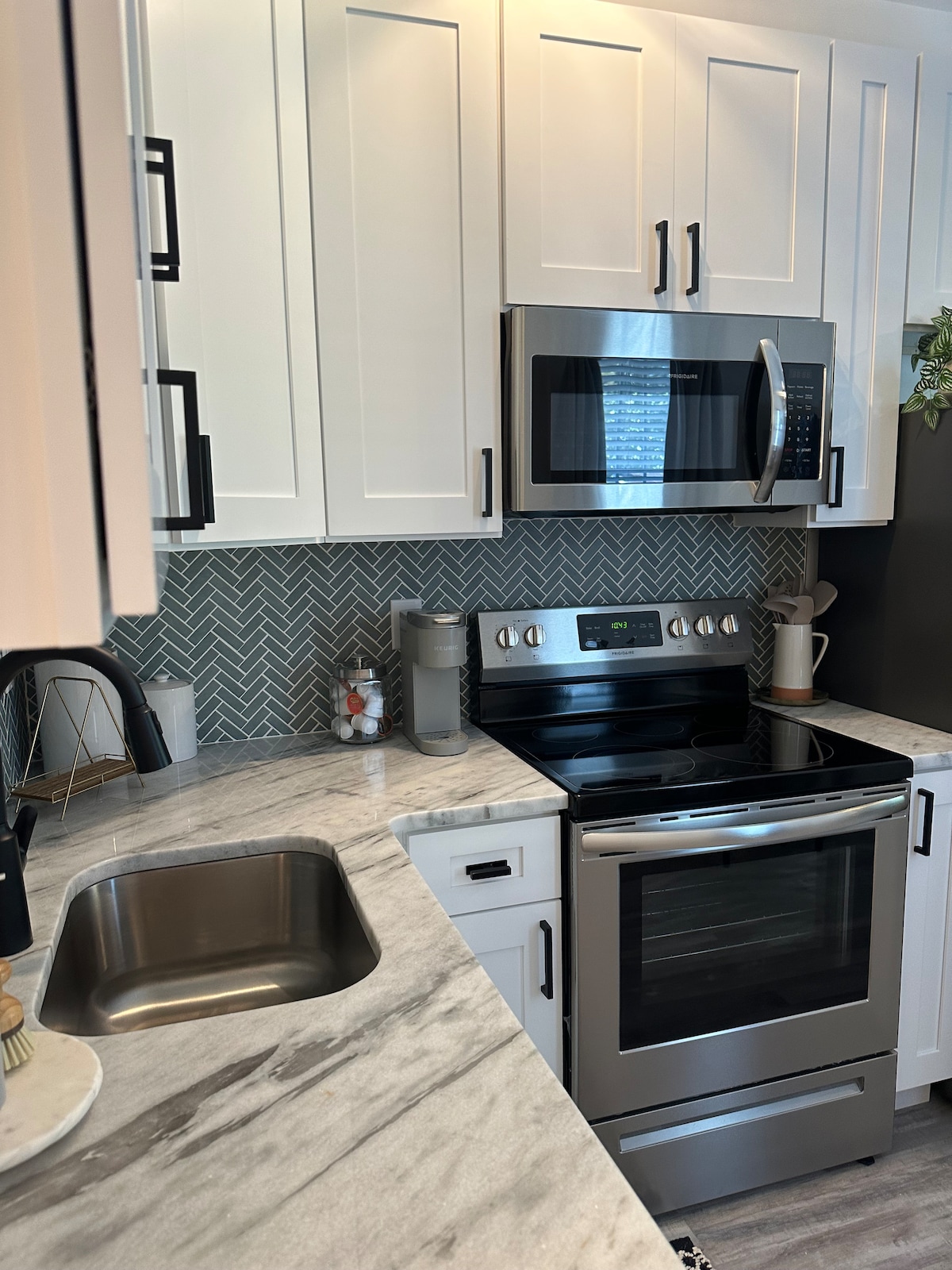 The kitchen showcases modern cabinetry with a sleek marble countertop. Stainless steel appliances, including an over-the-range microwave and an oven, are visible. A herringbone backsplash adds texture, while essential cookware is neatly organized on the counters.