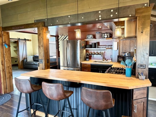 A modern kitchen showcases a wooden countertop with three bar stools positioned in front. Stainless steel appliances, including a refrigerator and gas stove, provide functional elegance. Shelving holds kitchenware and utensils, while ambient lighting adds warmth to the space.