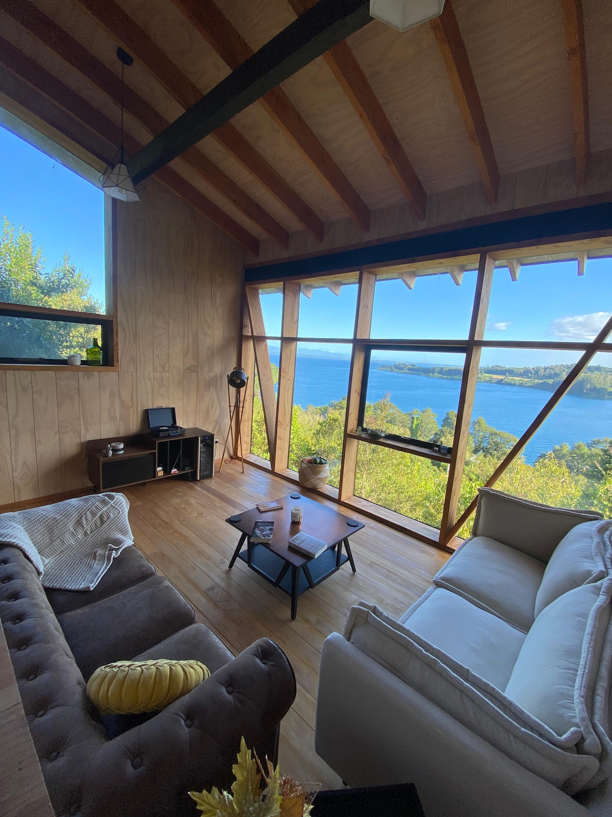 A cozy living area is highlighted by large windows that provide panoramic views of Lake Llanquihue. Two sofas are arranged around a central coffee table, while wooden walls enhance the natural ambiance. A small entertainment console and a decorative lamp add functional elements to the space.