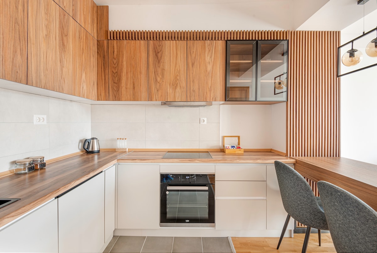 A modern kitchen features wooden cabinetry paired with sleek white units. An oven and stovetop are integrated into the countertop, complemented by a kettle and various kitchen accessories. Two charcoal grey chairs are positioned beside a wooden table, creating a functional dining area.