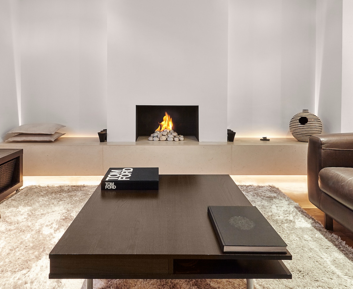 A minimalist living space features a low wooden coffee table positioned in front of a sleek fireplace, surrounded by smooth stones. Soft lighting highlights the textured area rug and two comfortable cushions placed on the floor. A modern vase adds elegance to the setting.