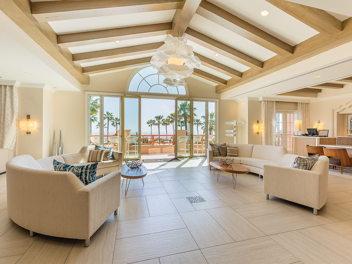 A spacious living area features soft furnishings arranged around a central table, with large glass doors providing natural light and views of palm trees outside. The high ceiling showcases wooden beams and a unique light fixture, enhancing the airy atmosphere of the room.