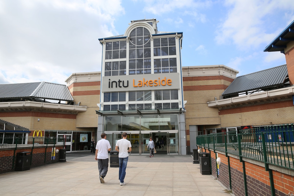 The entrance of intu Lakeside is shown, featuring large glass doors and a prominent signage. The structure’s architecture is modern, with a mix of brick and metal elements. A few people are walking towards the entrance, which is surrounded by outdoor seating areas.