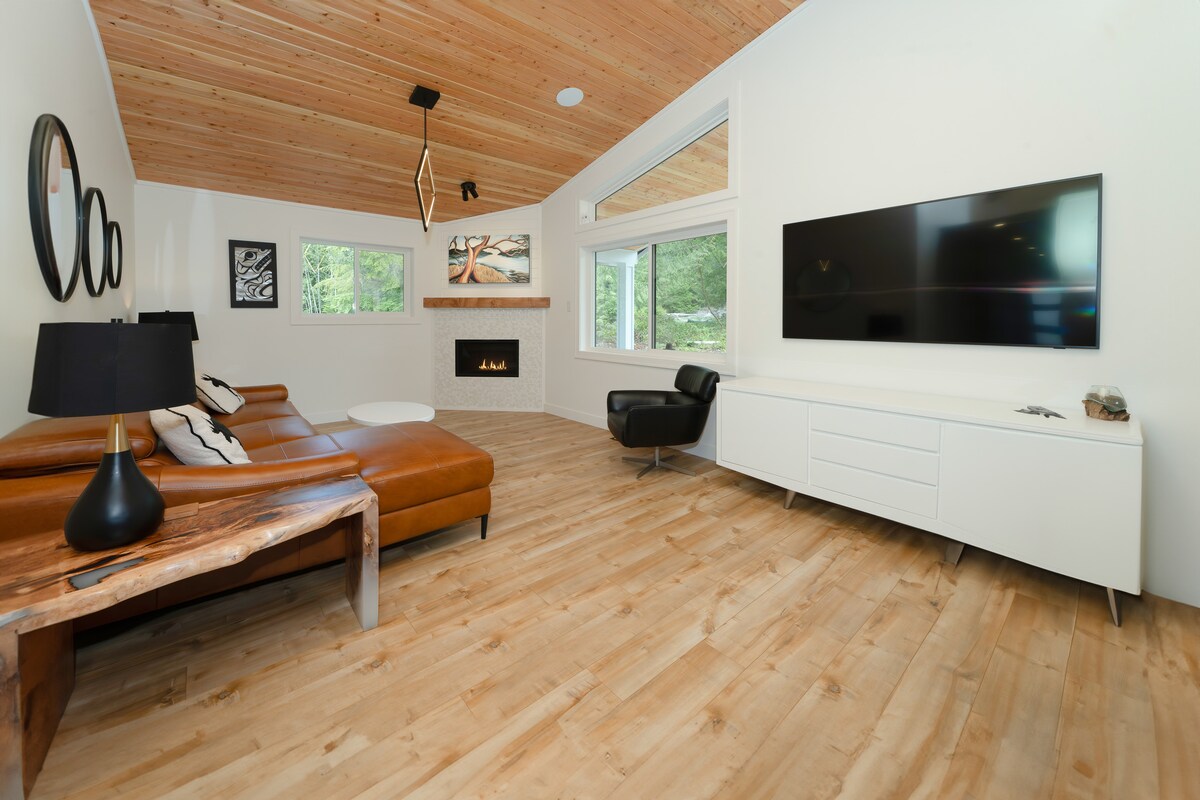 A modern living area is highlighted by a large sectional sofa and a sleek black armchair. Natural light fills the space through multiple windows. A recessed fireplace is centered on a white wall, complemented by wood accents on the ceiling and floor.