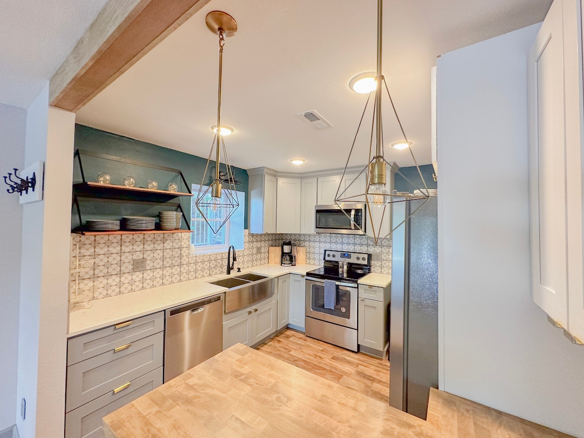The modern kitchen features elegant pendant lighting and a spacious countertop. Grey cabinetry and white appliances complement the intricate backsplash. A window provides natural light, while open shelving displays dishware, adding charm to the functional space.