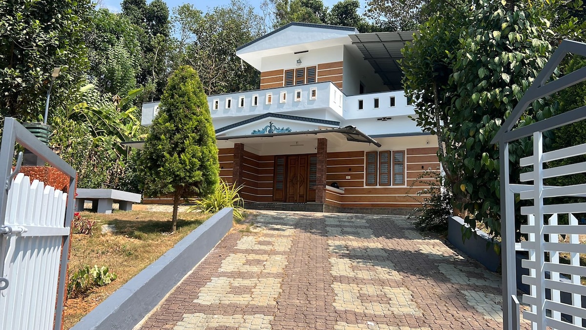 The exterior of a spacious home is framed by lush greenery. A wide pathway leads to the entrance, bordered by decorative stone paving. The two-story structure features multiple large windows and a welcoming porch. The front yard is landscaped with shrubs and a small tree.