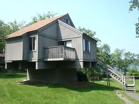 A rustic cabin is elevated on stilts, showcasing a gray exterior and a sloped roof. A small balcony with railing is visible, along with a stairway leading to the entrance. Surrounding greenery enhances the natural setting, with lake views in the distance.