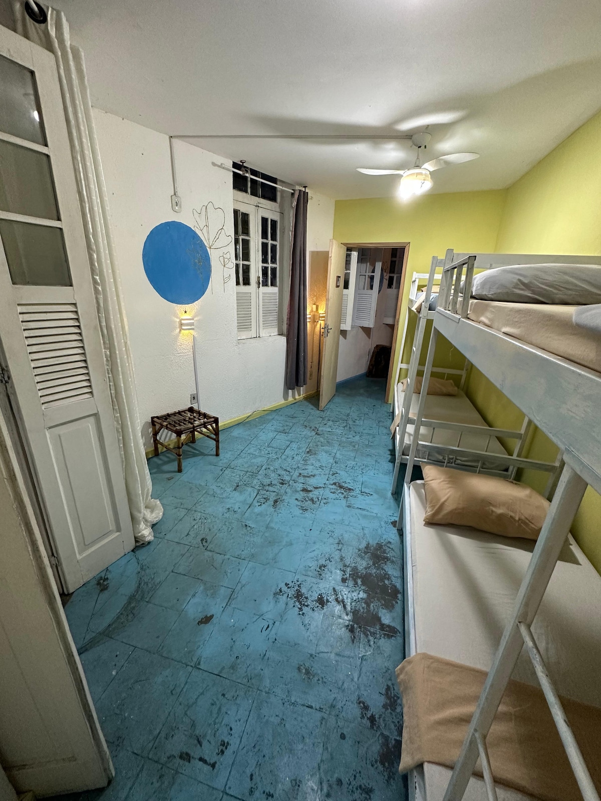 A bright room features two sets of bunk beds, providing accommodation for four guests. Walls are painted a soft yellow, while the floor is finished in a blue hue. Soft lighting is offered by wall fixtures, and a ceiling fan is visible above.