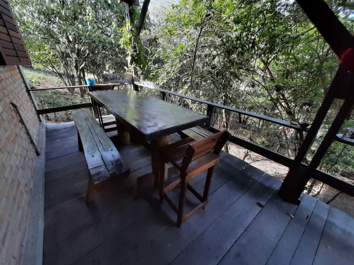 An outdoor dining area is featured under a covered balcony. A large wooden table is surrounded by bench seating and a chair, all set against a backdrop of lush greenery. Natural light filters through the trees, creating a calm and inviting space.
