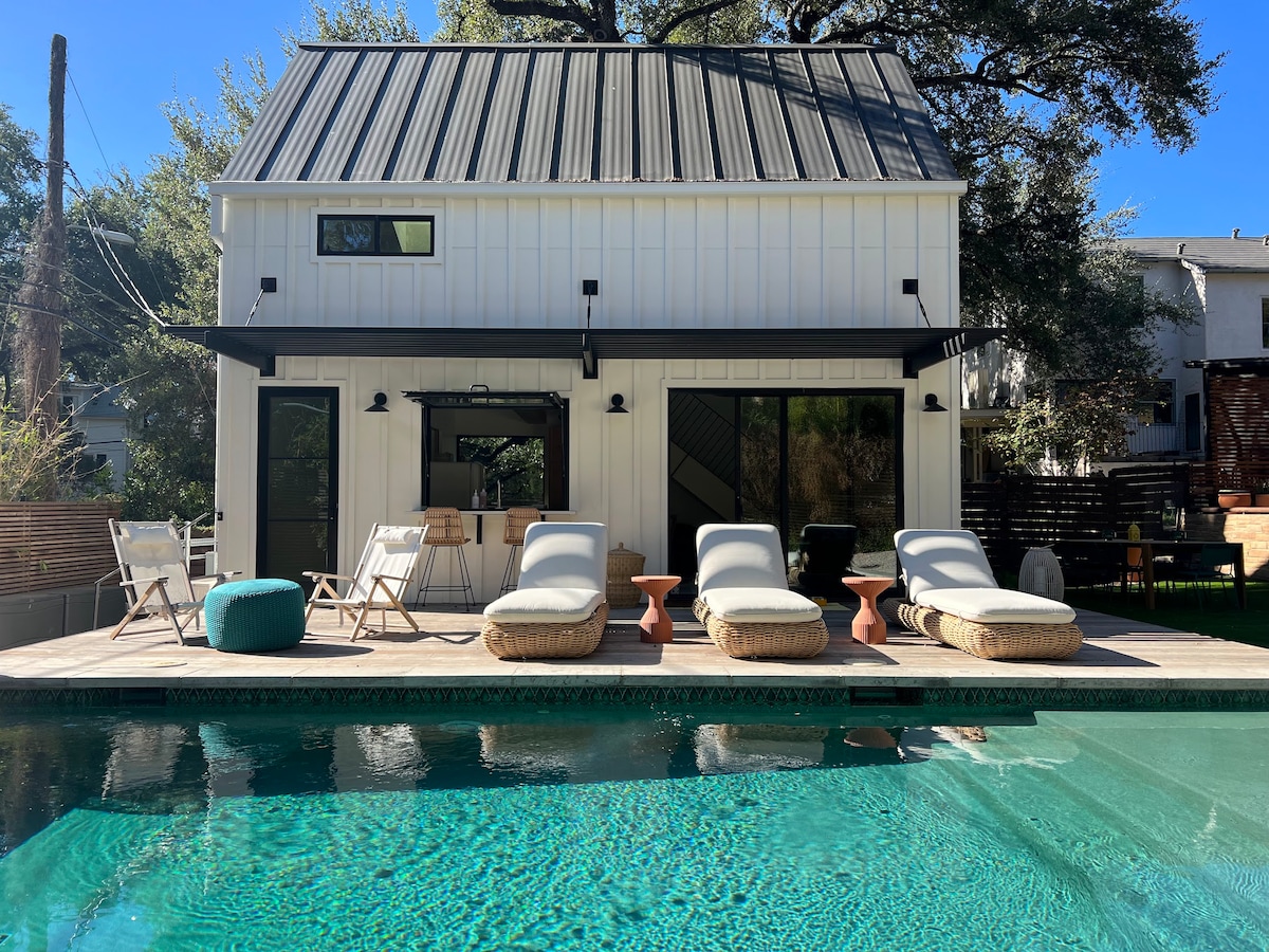 A modern casita is viewed from the poolside, featuring large windows and a flat metal roof. Four lounge chairs are positioned around a turquoise swimming pool, with a small table nearby. Lush greenery surrounds the outdoor area, providing a serene environment.