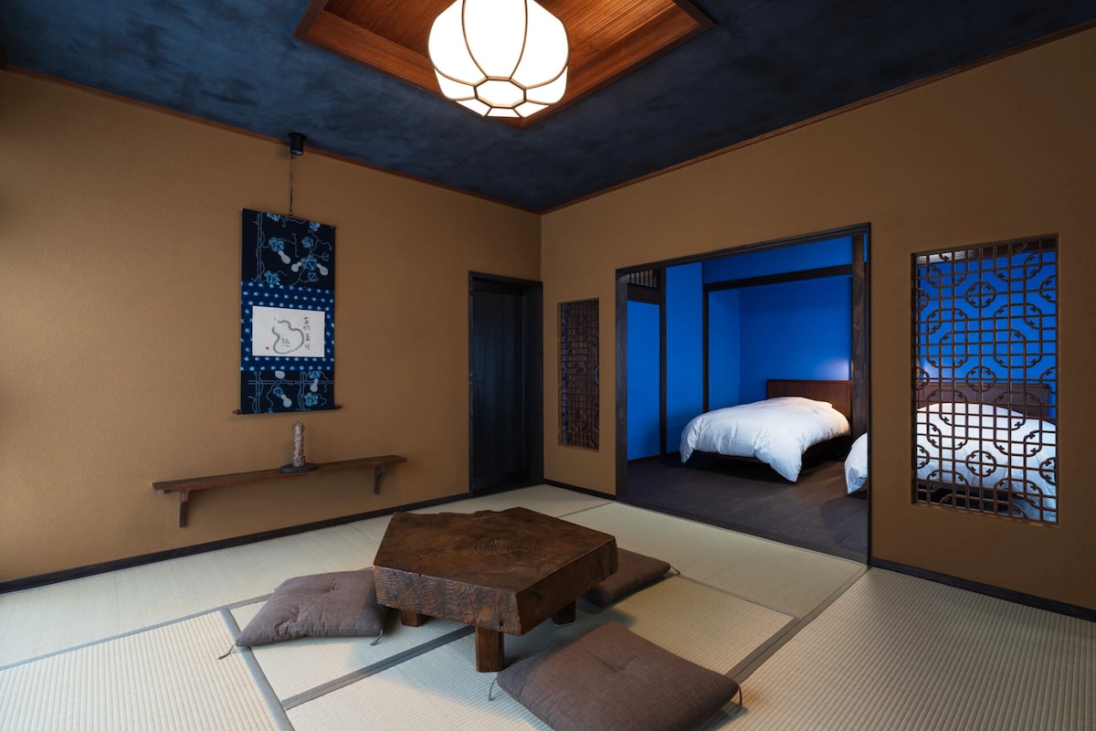 A spacious room features traditional tatami flooring and a wooden low table at the center, surrounded by floor cushions. Two doors lead to a blue-themed sleeping area, while decorative wall elements enhance the room's character.