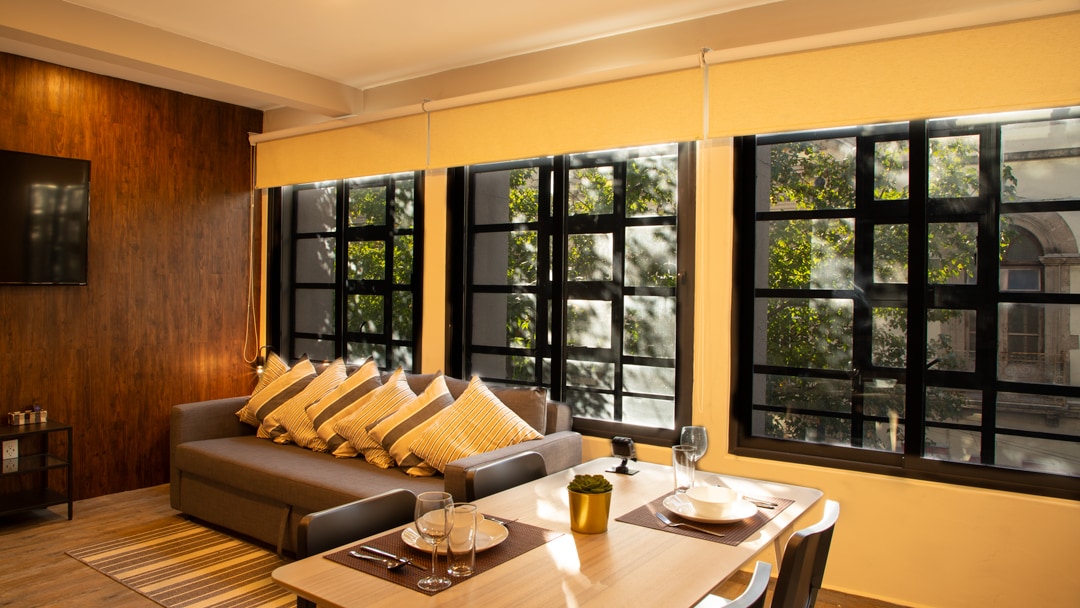 A cozy living area showcases a double sofa bed adorned with multiple decorative pillows. Large black-framed windows allow natural light to illuminate the space, with greenery visible outside. A dining table is set with glassware and a small plant, completing the inviting atmosphere.