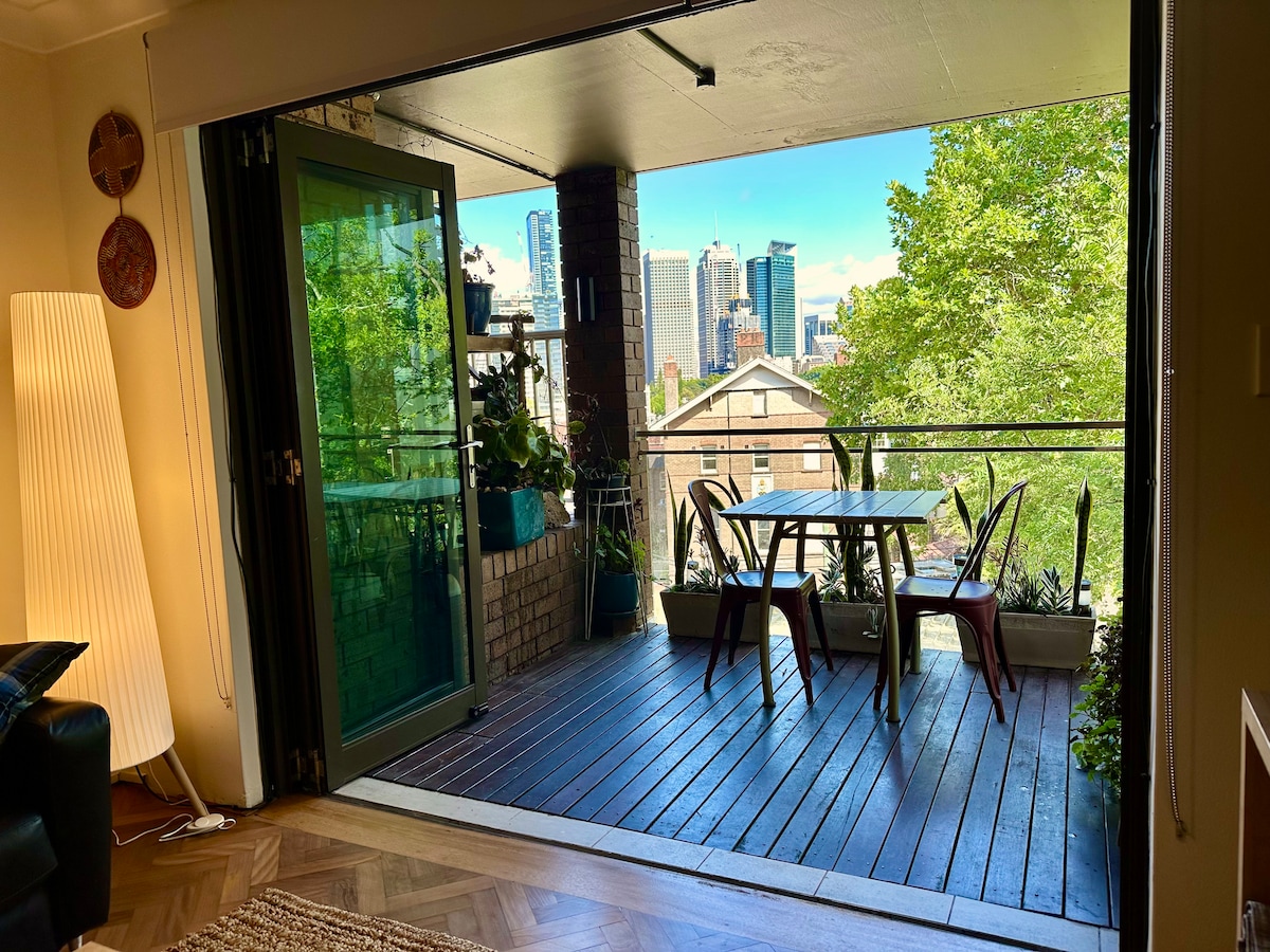 The image depicts a balcony with a wooden deck featuring a table and four chairs, surrounded by greenery. Large glass doors open to showcase city views under a clear sky. The living area is partially visible, connecting indoor and outdoor spaces.