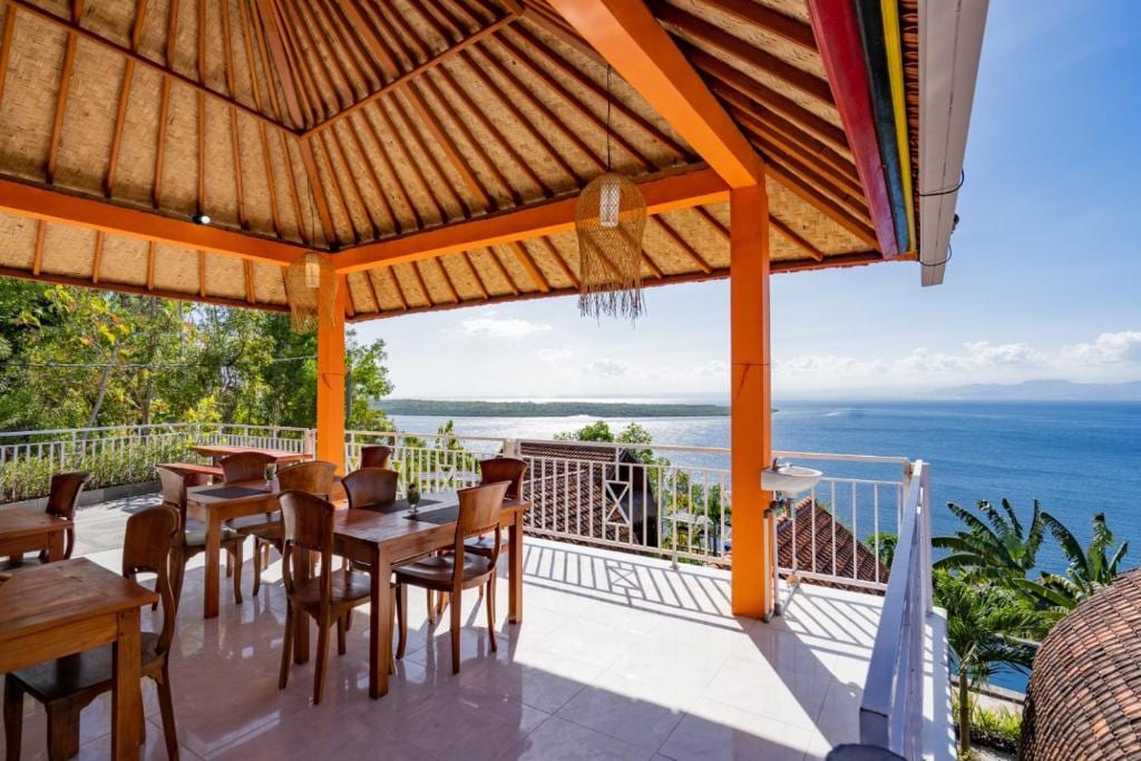 An open dining area features wooden tables and chairs under a high thatched roof. The space offers expansive views of the ocean and surrounding greenery, with bright orange accents enhancing the vibrant atmosphere. Natural light floods the area, creating a welcoming environment.