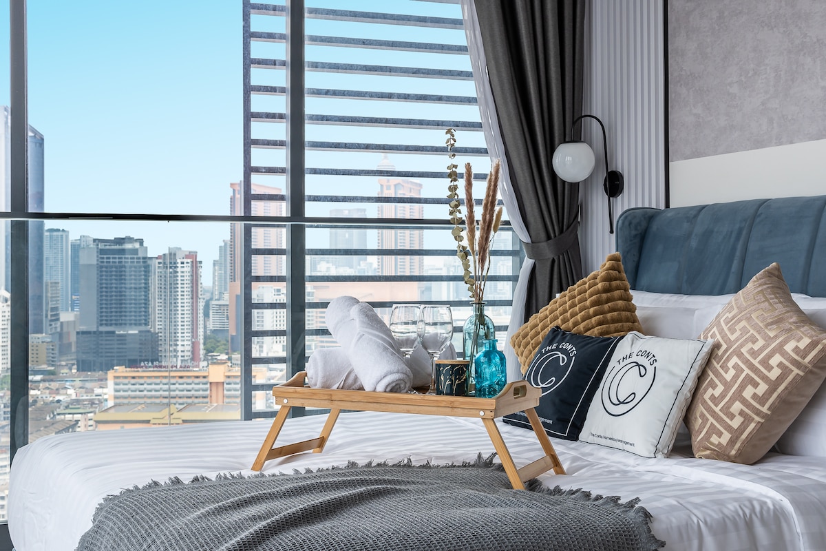 A spacious bed is presented with neatly arranged towels and decorative pillows. A tray with glassware rests on the bed, while large windows offer a view of the city skyline, allowing light to brighten the room. Soft furnishings enhance the overall comfort.