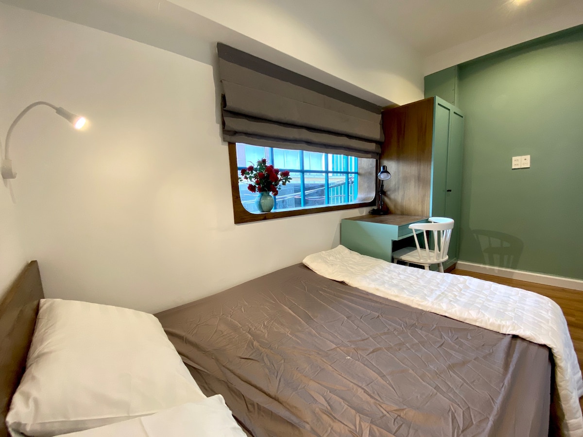 A well-lit sleeping area features a spacious bed dressed in neutral bedding, with two white pillows. A small table and chair are positioned near a window adorned with a decorative vase of flowers. Soft green walls enhance the overall calming ambiance.