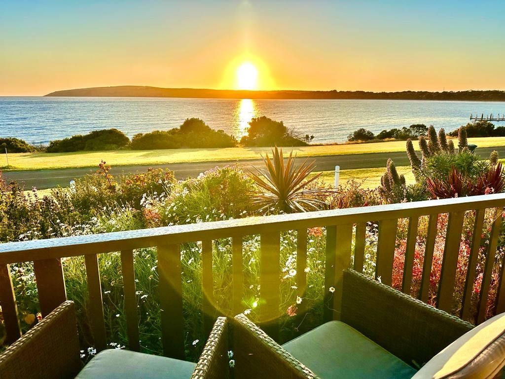 The front deck features comfortable seating with a view of the sunrise over the ocean. Lush gardens filled with various plants and flowers lead to the waterfront. A tranquil atmosphere is created as sunlight reflects off the water, enhancing the coastal scenery.