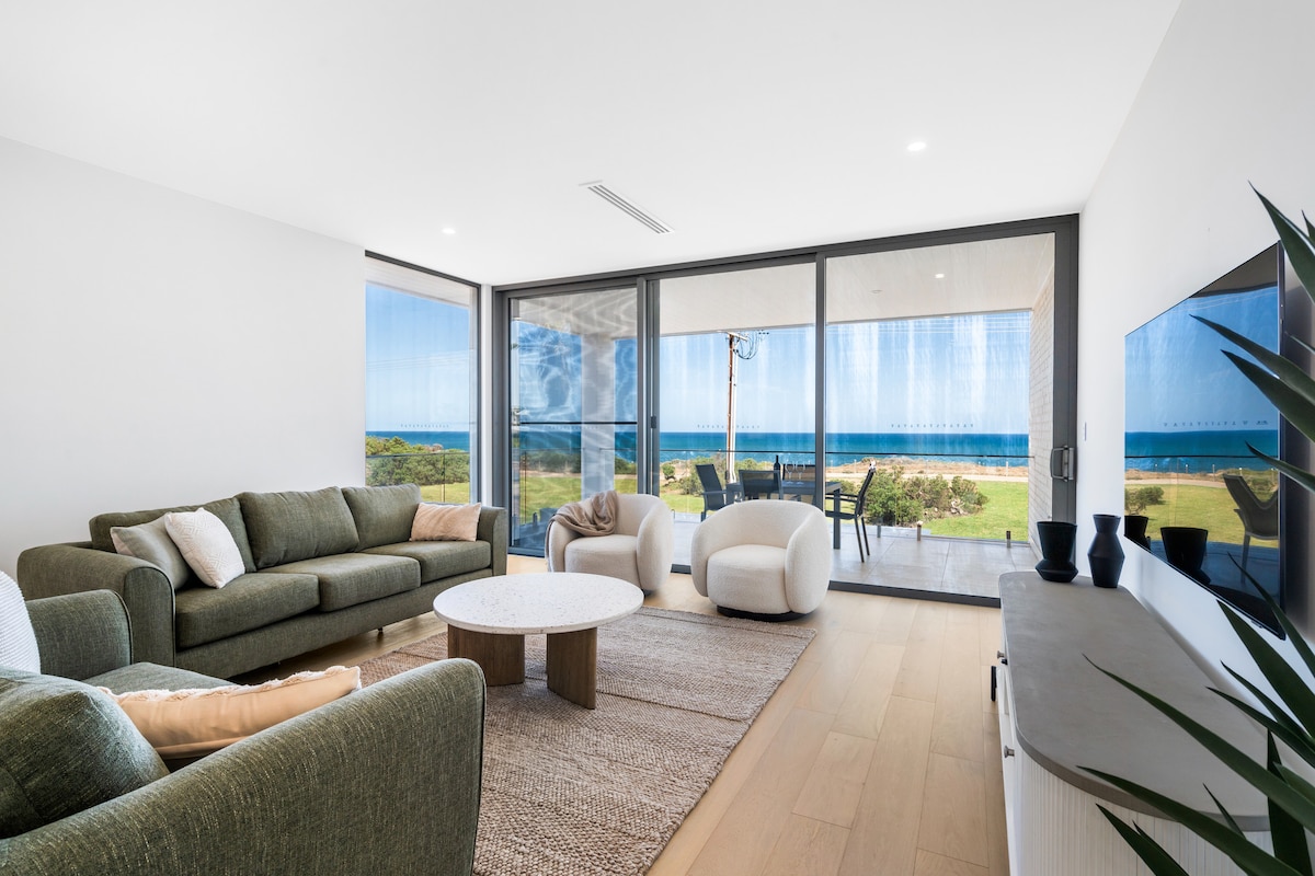 The spacious living area features a contemporary design with large glass sliding doors that offer panoramic ocean views. Comfortable seating is arranged around a circular coffee table, enhancing the inviting atmosphere. Natural light floods the room, complemented by neutral tones and a textured rug.