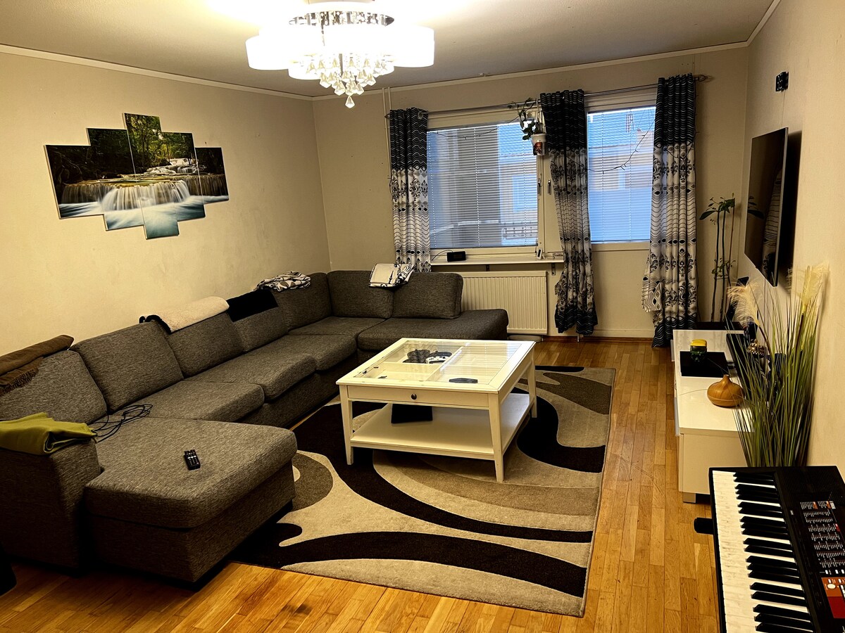 A spacious living area features a large gray sectional sofa arranged around a central white coffee table. A patterned area rug lies beneath, complementing the wooden flooring. Dramatic curtains frame the window, while a television and a keyboard are positioned against the wall.