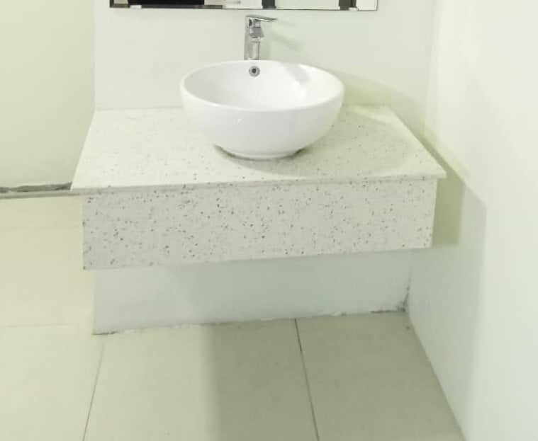 A clean and modern bathroom sink area is displayed, featuring a round, white vessel sink set on a minimalist speckled countertop. The wall behind the sink is plain, and a mirror is mounted above, enhancing the sense of openness.
