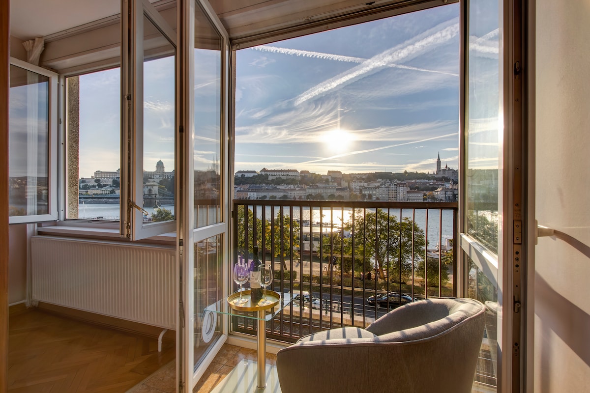 A view from the balcony offers a glimpse of the Danube River and the sunset reflecting on the water. Two glass doors open to the outside, allowing natural light to fill the space. A small table and chair are positioned on the balcony, enhancing the scenic experience.