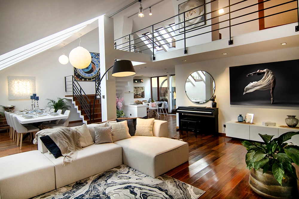 A spacious living area is highlighted by a modern sectional sofa and a large area rug with abstract patterns. A grand piano sits nearby, while a dining table for eight is set in the background. Natural light pours in through the ceiling windows, enhancing the elegant decor.