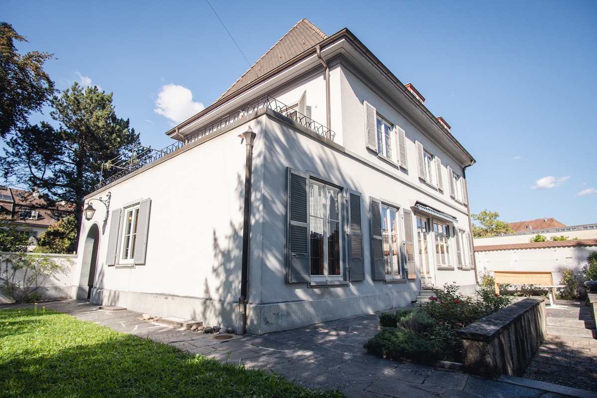 The villa features a light-colored exterior with large windows that allow natural light to fill the rooms. A well-maintained garden surrounds the property, adding greenery. The roof has a slight overhang, and the structure showcases a blend of classic architectural elements.
