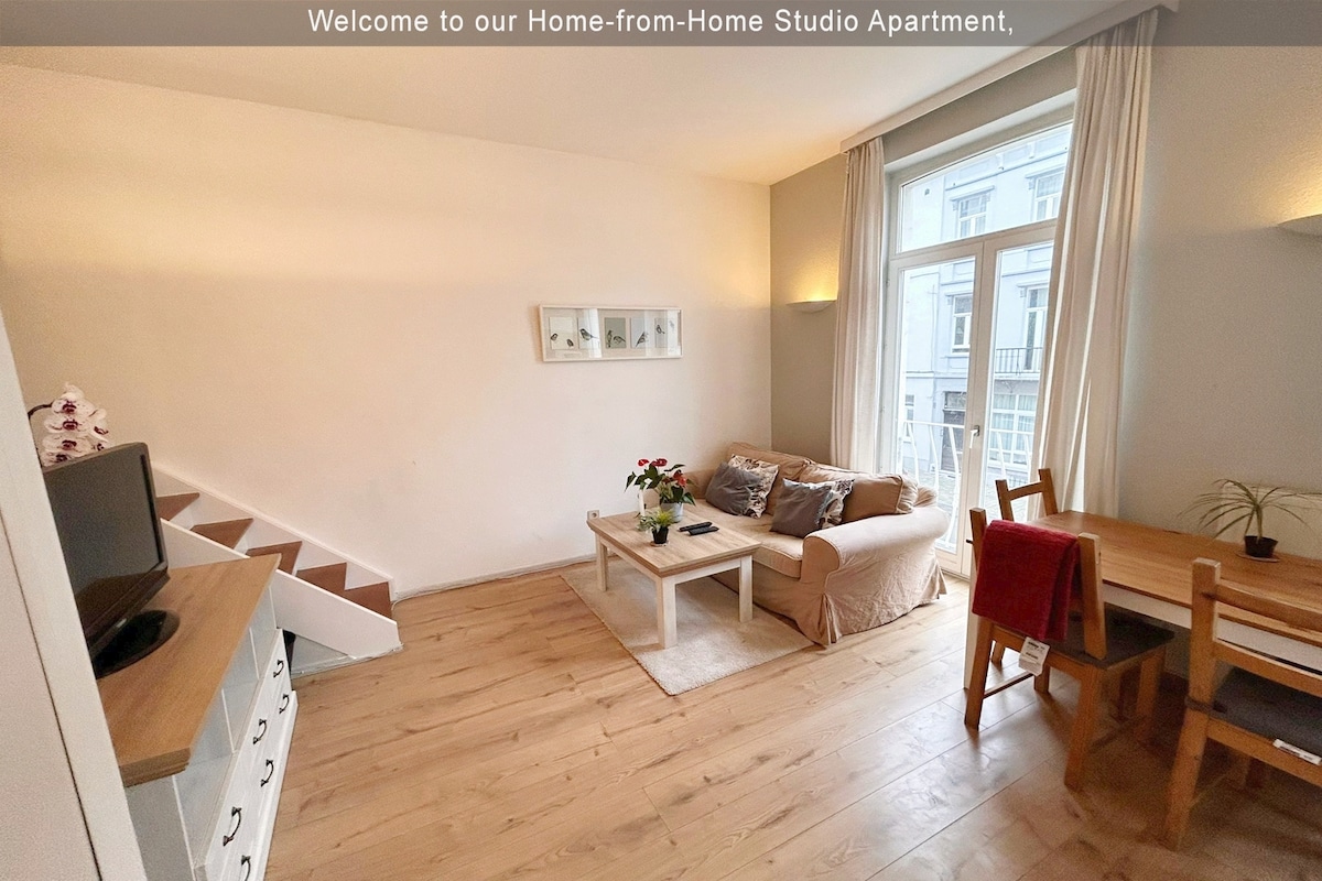 The bright living room features large windows that provide natural light, a comfortable two-seater sofa beside a small coffee table, and a dining area with a table for two. Light wooden flooring complements the warm and welcoming decor.