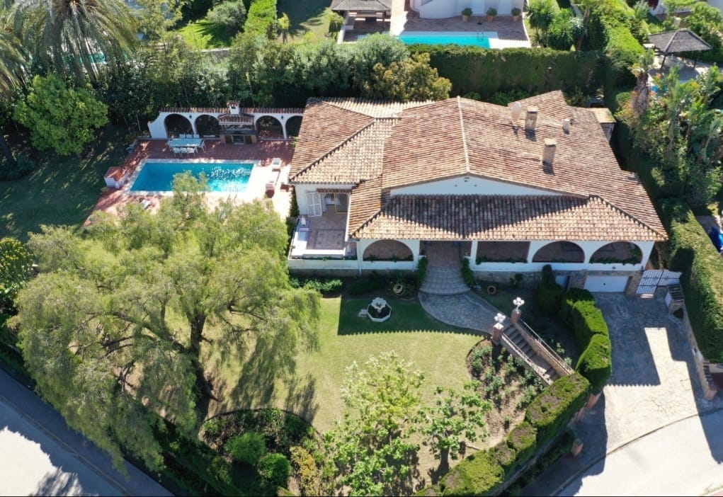 An aerial view captures the unique villa surrounded by lush greenery. It features a spacious pool area with loungers and a manicured garden, enhancing the serene outdoor environment. The villa's terracotta roof complements the classic Andalusian architecture, with a clear pathway leading towards the entrance.