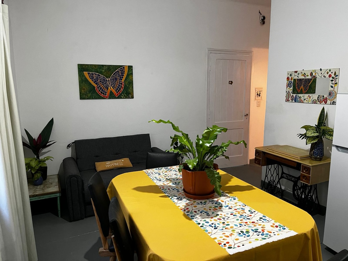 A dining area featuring a yellow tablecloth laid over a rectangular table is highlighted by a central potted plant. Surrounding chairs are arranged for shared meals. A bright butterfly artwork adorns the wall, while a cozy gray sofa and a decorated mirror complement the inviting space.
