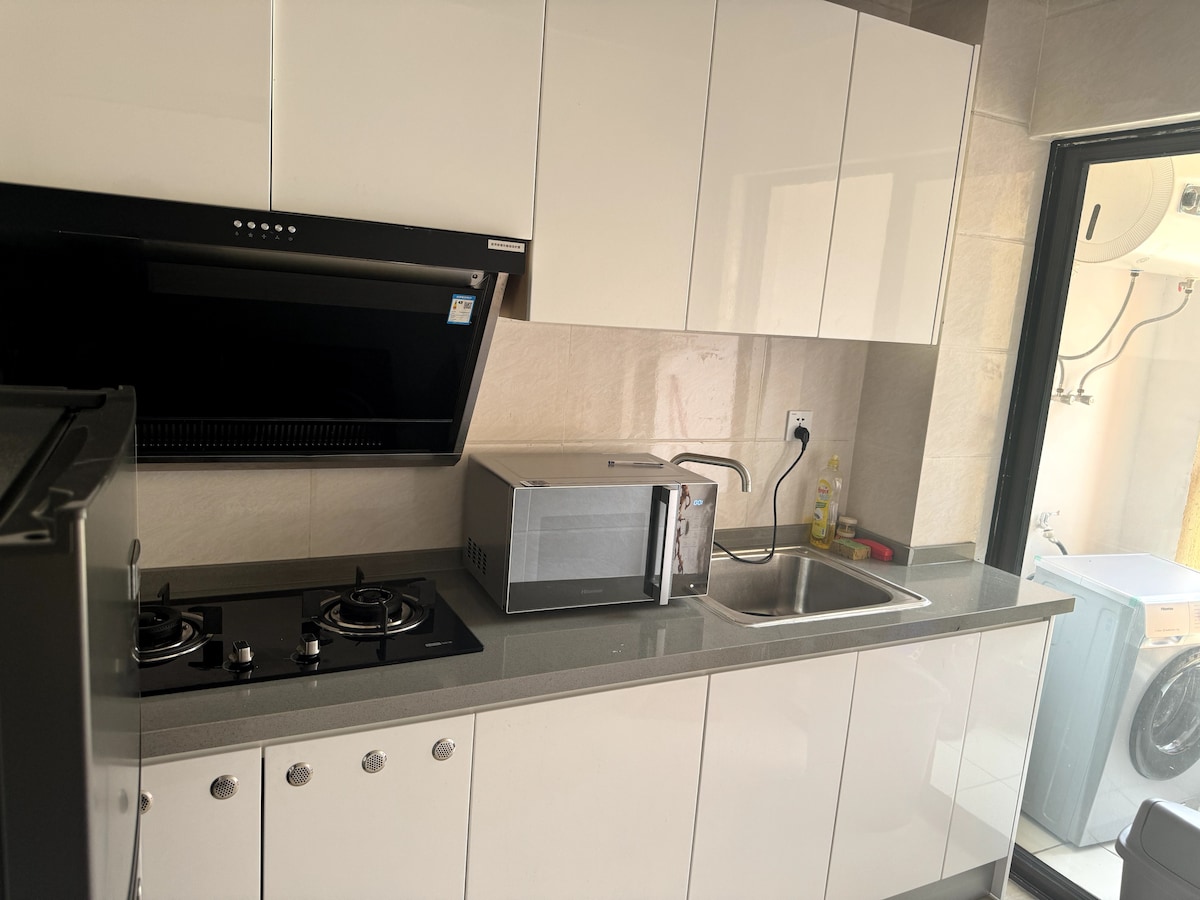 A modern kitchen is presented with sleek white cabinetry and a gray countertop. Cooking appliances include a gas stove with two burners and a microwave. A stainless steel sink is visible, and natural light enters through a nearby door leading to an outdoor area.