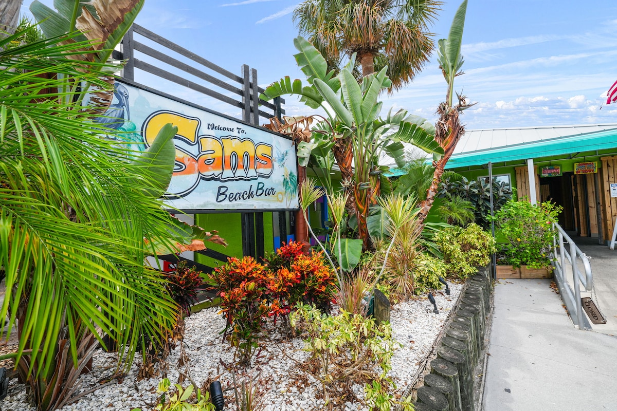 A colorful entrance to Sam's Beach Bar is surrounded by lush tropical plants, including palms and flowering shrubs. The sign displays the establishment's name, providing a welcoming atmosphere. The pathway leading to the entrance is lined with well-maintained landscaping, creating an inviting approach.