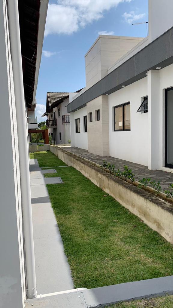A narrow pathway is lined with grass and bordered by two white structures, showcasing modern architectural design. The building on the right features large windows and a sleek facade, while the left side presents a mix of traditional elements with balconies visible in the distance.