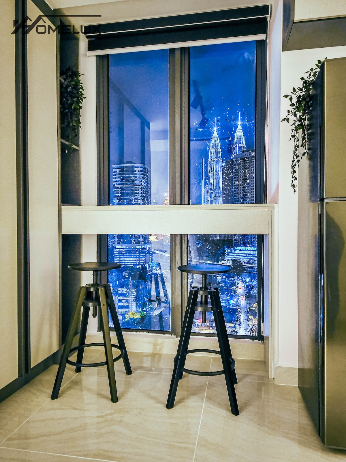 A kitchen area features two black stools positioned at a window overlooking the Kuala Lumpur skyline. The iconic Petronas Twin Towers are clearly visible against the evening sky, providing a stunning urban backdrop. Soft lighting highlights the modern cabinetry and countertop.
