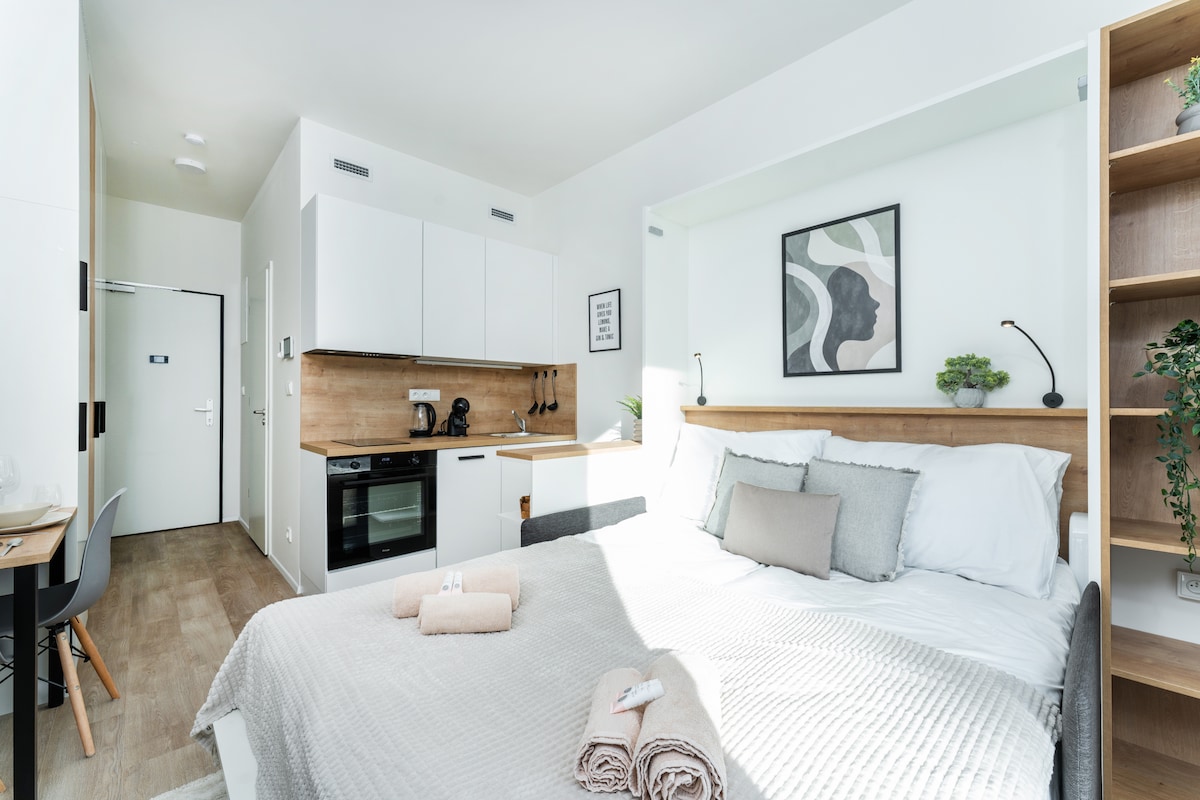 A modern studio apartment features a spacious bed adorned with neutral-colored bedding. A kitchenette with minimalistic cabinetry and essential appliances is visible in the background. Natural light floods the room through large windows, creating a bright and airy ambiance. Decorative plants add a touch of greenery.