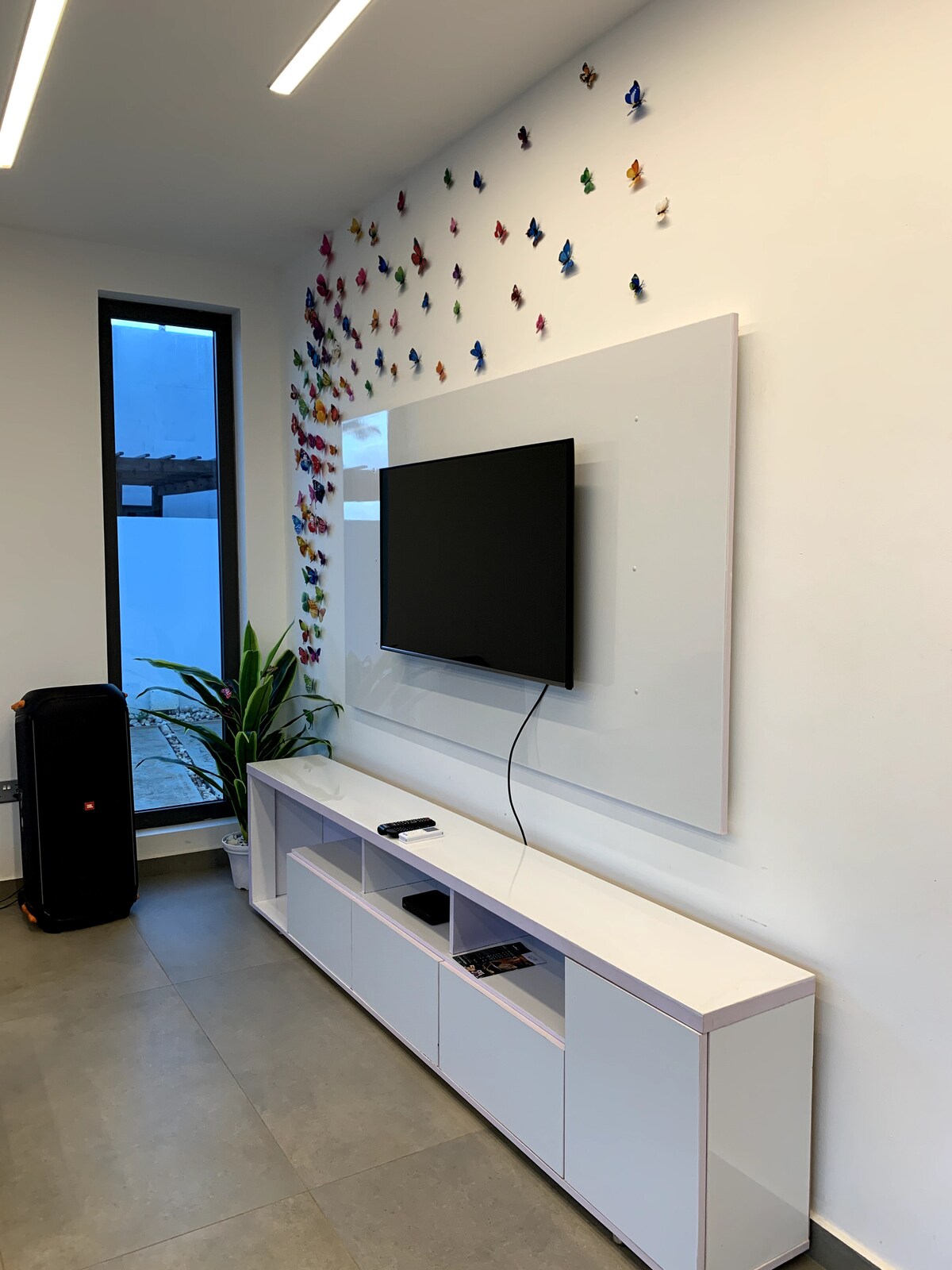 A modern living area features a sleek entertainment unit with a mounted flat-screen television. Colorful butterfly decorations adorn the white wall above, while a tall plant adds a touch of greenery beside a large window that allows natural light to enter the space.